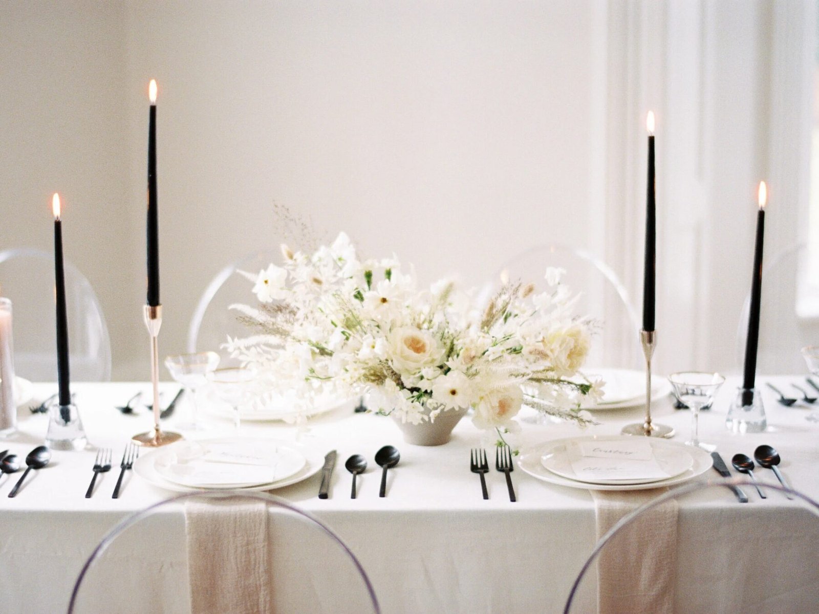 Editorial tablescape with low ivory-and-dried centerpiece, tall black tapers, ghost chairs and blush runner against a bright neutral room