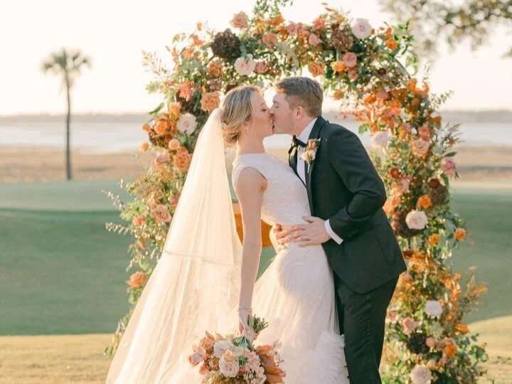 Couple kissing under a lush autumnal floral arch in rust, peach and blush tones with a waterfront backdrop at golden hour