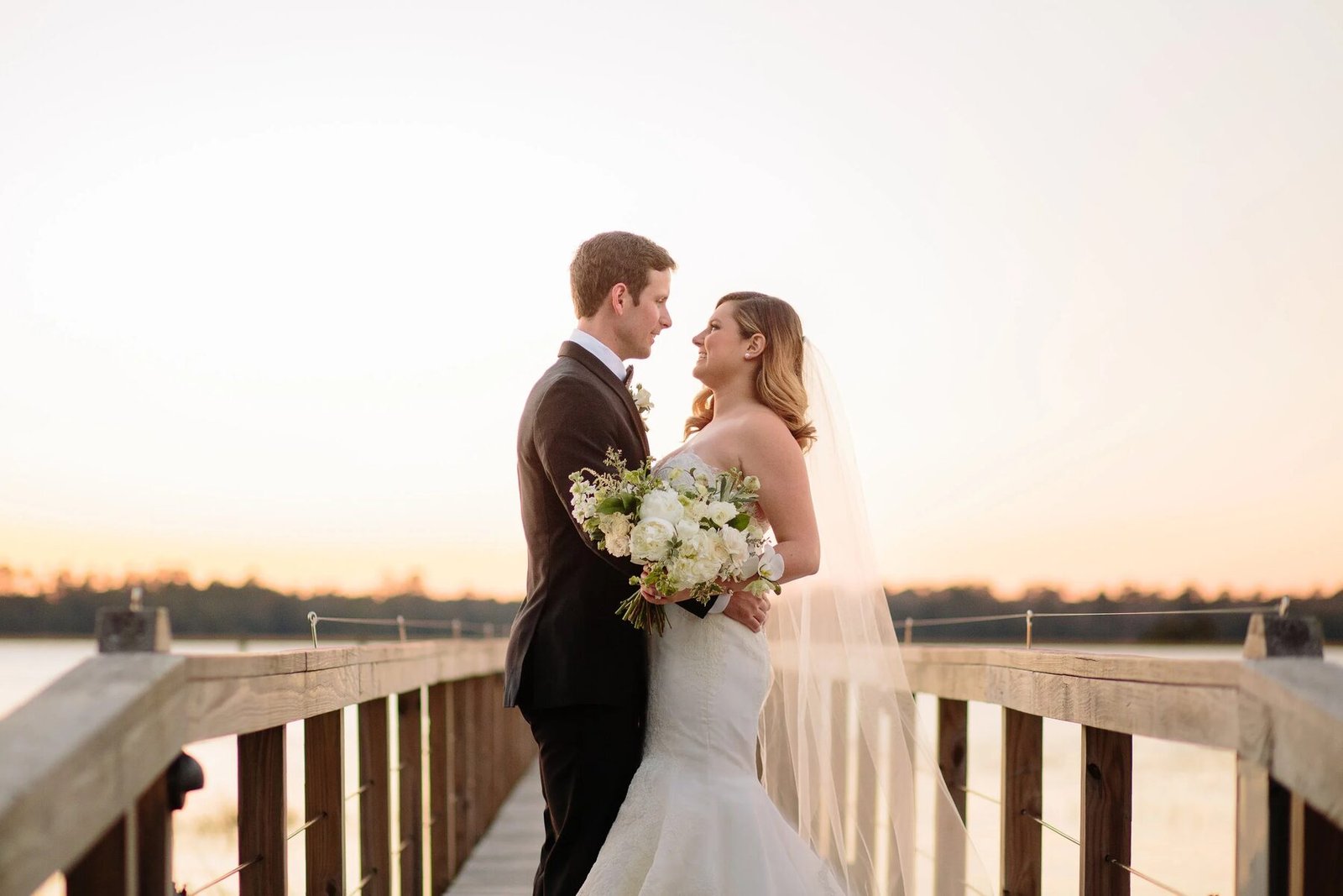 Bride and groom embracing on a wooden dock at golden-hour sunset over the water, bride holding ivory bouquet