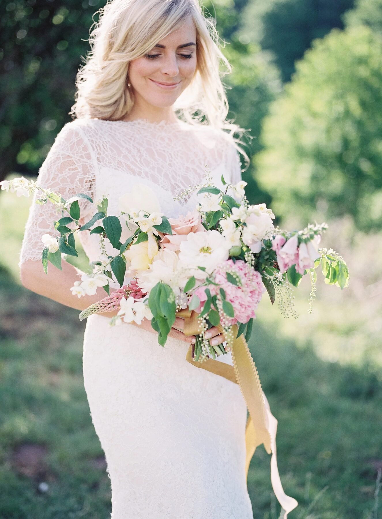 Backlit bride in lace overlay gown cradling soft pink/cream bouquet, sunlit greenery behind