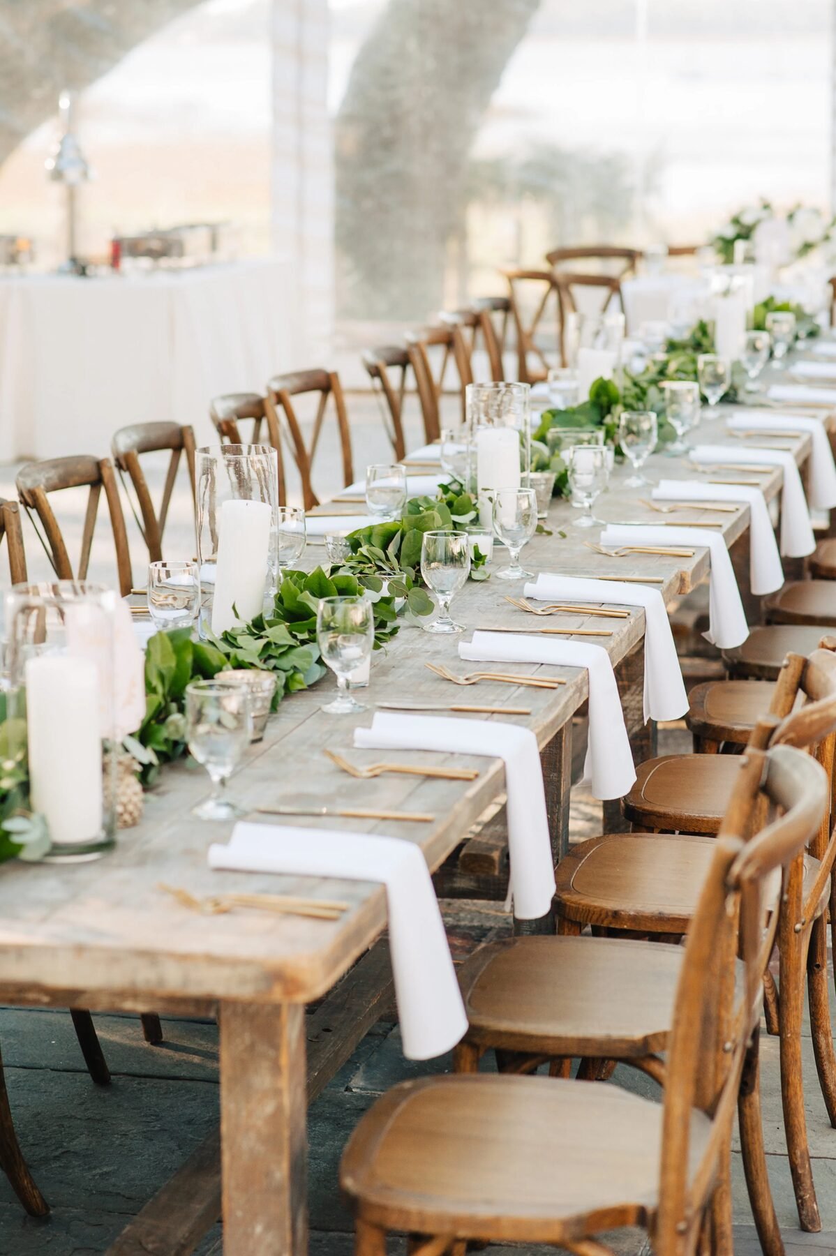 Long wood farm table with lush green garland runner, white taper candles and folded napkins under a tent