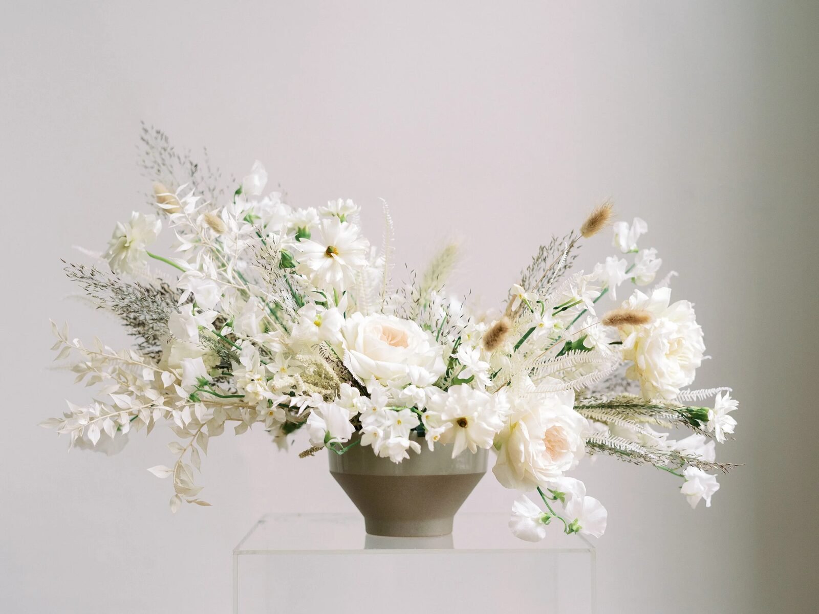 All-ivory garden arrangement (roses, cosmos, dried grasses, bunny tail) in a stone compote on an acrylic plinth against a soft neutral wall